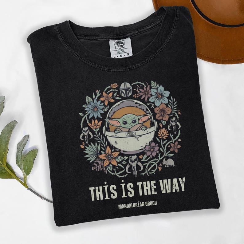 May include: Black t-shirt featuring a floral wreath design with a cartoon image of Grogu from The Mandalorian. The text "THIS IS THE WAY" and "MANDALORIAN GROGU" are printed below the image. The shirt is a Comfort Colors brand.
