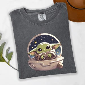Puede incluir: Camiseta gris oscuro con una imagen de dibujos animados de un personaje alien&iacute;gena verde en una c&aacute;psula contra un fondo estrellado. La camiseta tiene cuello redondo y mangas cortas. La etiqueta dice "Comfort Colors".
