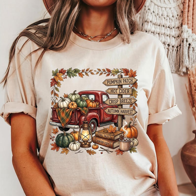Puede incluir: Una camiseta beige con un gr&aacute;fico de una camioneta roja cargada de calabazas, una linterna y libros. Un poste de madera muestra el texto PUMPKIN PATCH, HOT CIDER, CRISP BREEZE y AUTUMN HARVEST, rodeado de hojas de oto&ntilde;o.