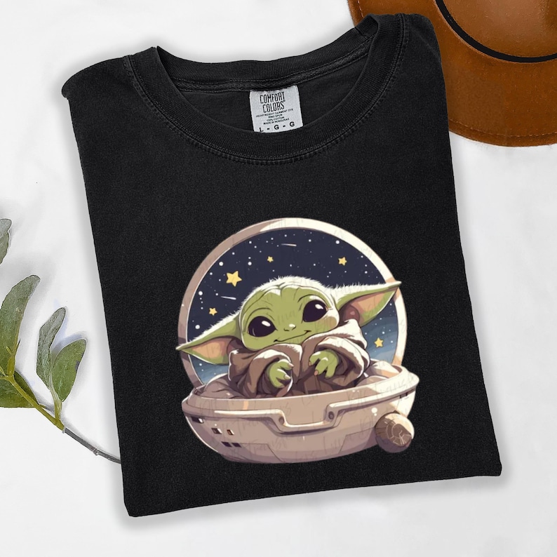 Puede incluir: Camiseta negra con una ilustraci&oacute;n de dibujos animados de un personaje verde alien&iacute;gena en una c&aacute;psula espacial sobre un fondo estrellado. La camiseta tiene cuello redondo y la etiqueta de la marca Comfort Colors.