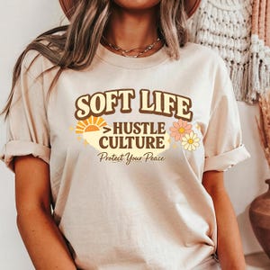 May include: Beige t-shirt with the text "SOFT LIFE > HUSTLE CULTURE" in brown and yellow, with a sun and flower graphic. The phrase "Protect Your Peace" is printed below in a script font. The shirt has rolled-up sleeves.