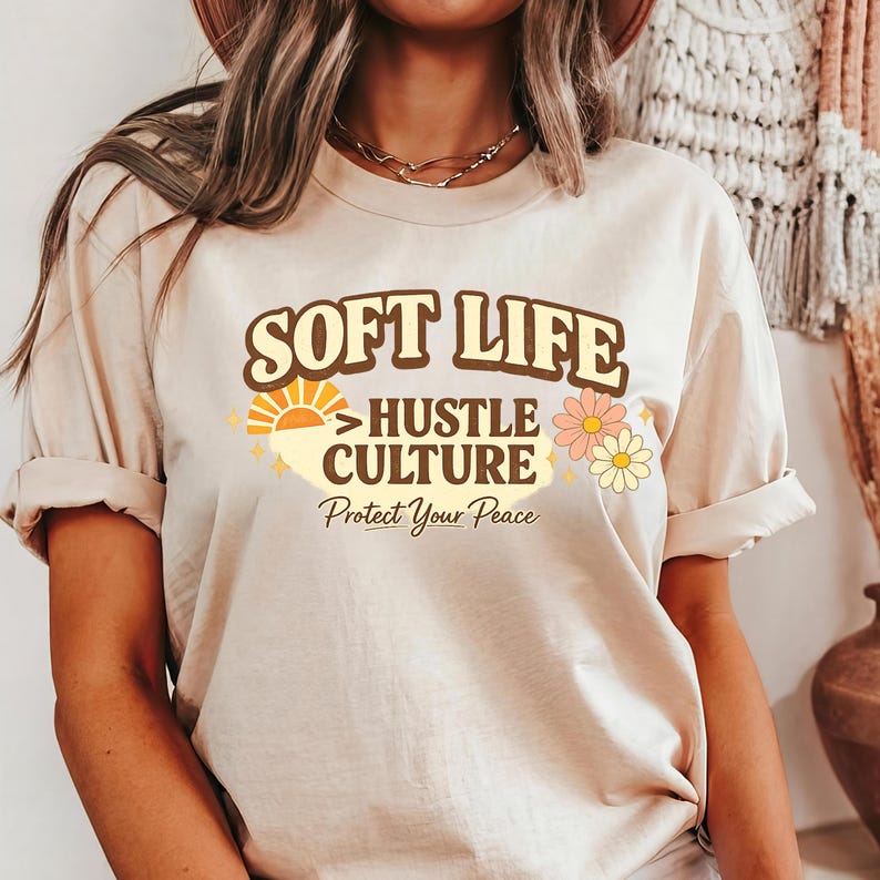 May include: Beige t-shirt with the text "SOFT LIFE > HUSTLE CULTURE" in brown and yellow, with a sun and flower graphic. The phrase "Protect Your Peace" is printed below in a script font. The shirt has rolled-up sleeves.