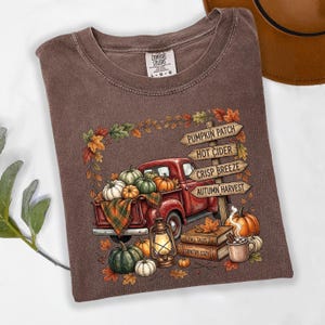 Puede incluir: Una camiseta marr&oacute;n de cuello redondo con un gr&aacute;fico de una camioneta roja cargada de calabazas, una linterna y libros. Un poste de madera muestra el texto: PUMPKIN PATCH, HOT CIDER, CRISP BREEZE, AUTUMN HARVEST, FALL TALES, COUNTRY COZY.