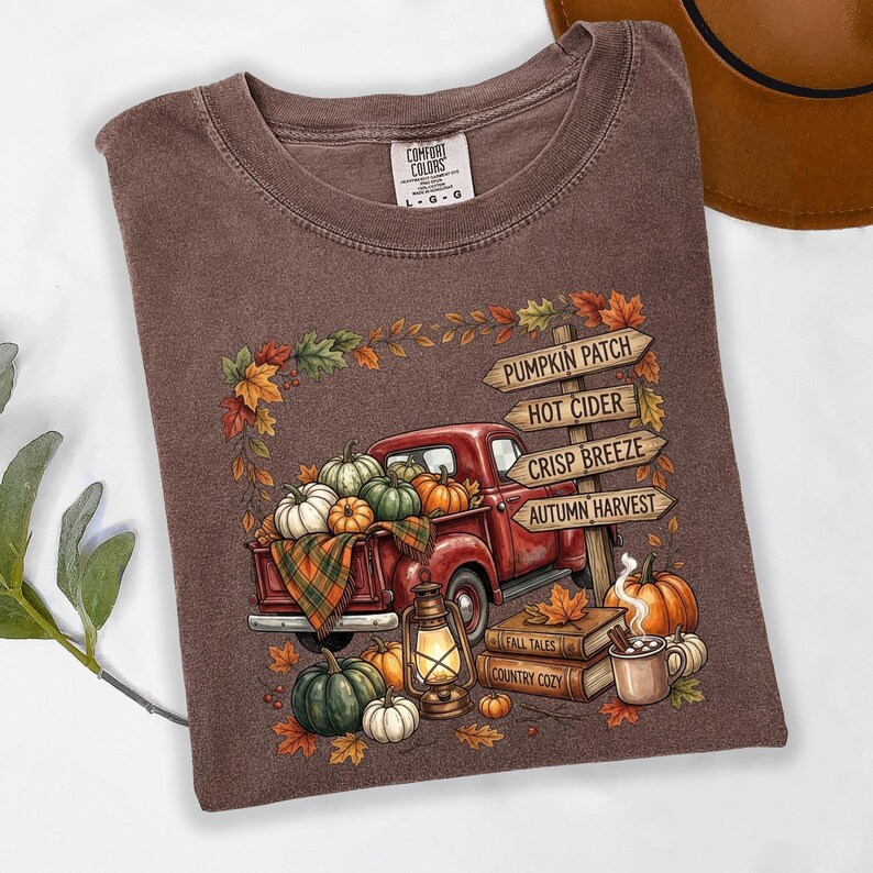 Puede incluir: Una camiseta marr&oacute;n de cuello redondo con un gr&aacute;fico de una camioneta roja cargada de calabazas, una linterna y libros. Un poste de madera muestra el texto: PUMPKIN PATCH, HOT CIDER, CRISP BREEZE, AUTUMN HARVEST, FALL TALES, COUNTRY COZY.