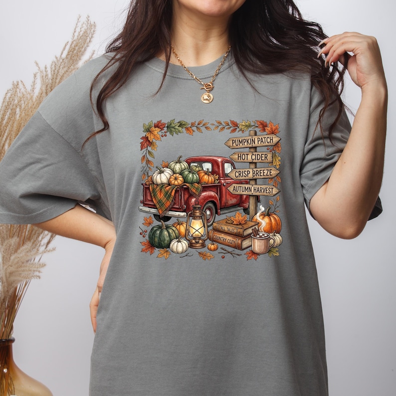Puede incluir: Una camiseta gris con un gr&aacute;fico de una camioneta roja llena de calabazas, una linterna y libros. Un poste de madera muestra el texto PUMPKIN PATCH, HOT CIDER, CRISP BREEZE y AUTUMN HARVEST, rodeado de hojas de oto&ntilde;o.