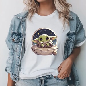 Puede incluir: Camiseta blanca con una imagen de dibujos animados de un personaje alien&iacute;gena verde en una c&aacute;psula espacial sobre un fondo estrellado. El extraterrestre tiene orejas grandes y ojos grandes. La camiseta se usa con una chaqueta vaquera.