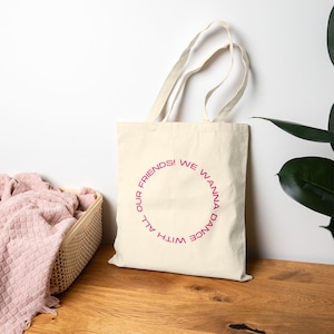 Puede incluir: Bolso tote de color crema con el texto rosa "WE WANNA DANCE WITH ALL OUR FRIENDS!" en un diseño circular. El bolso tiene dos asas y está hecho de un material similar a la lona. El bolso está sobre una superficie de madera.