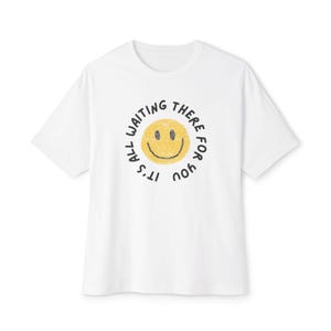 Puede incluir: Camiseta blanca con un diseño de cara sonriente amarilla en el centro. La cara sonriente está rodeada por el texto negro "It's all waiting there for you". La camiseta tiene mangas cortas y cuello redondo.