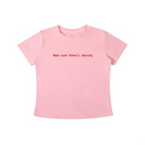 Könnte beinhalten: Ein hellrosa T-Shirt mit kurzen Ärmeln und Rundhalsausschnitt. In der Mitte der Brust befindet sich der Text "Make sure there's dancing", der in einer kleinen, roten, serifenlosen Schrift gedruckt ist. Das Kleidungsstück hat einen geraden Saum.