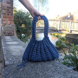 May include: A handmade blue knitted handbag with a braided handle. The bag is held by a person's hand, showcasing its unique design and texture. The bag has a flared shape and a tassel detail at the bottom.