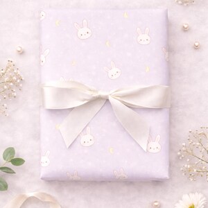 May include: A wrapped gift with light purple bunny-themed wrapping paper, tied with a white satin bow. The paper features repeating bunny faces, moons, and stars. The words "Before the Ribbon" are printed on the bottom left.
