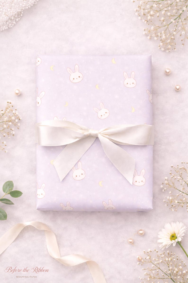 May include: A wrapped gift with light purple bunny-themed wrapping paper, tied with a white satin bow. The paper features repeating bunny faces, moons, and stars. The words "Before the Ribbon" are printed on the bottom left.
