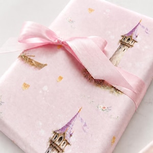 Puede incluir: Un regalo rectangular envuelto en papel rosa claro con un patr&oacute;n repetitivo de torres de piedra altas con techos c&oacute;nicos morados, botes peque&ntilde;os y detalles florales. Una cinta de sat&eacute;n rosa transl&uacute;cida est&aacute; atada en un lazo en la parte superior.