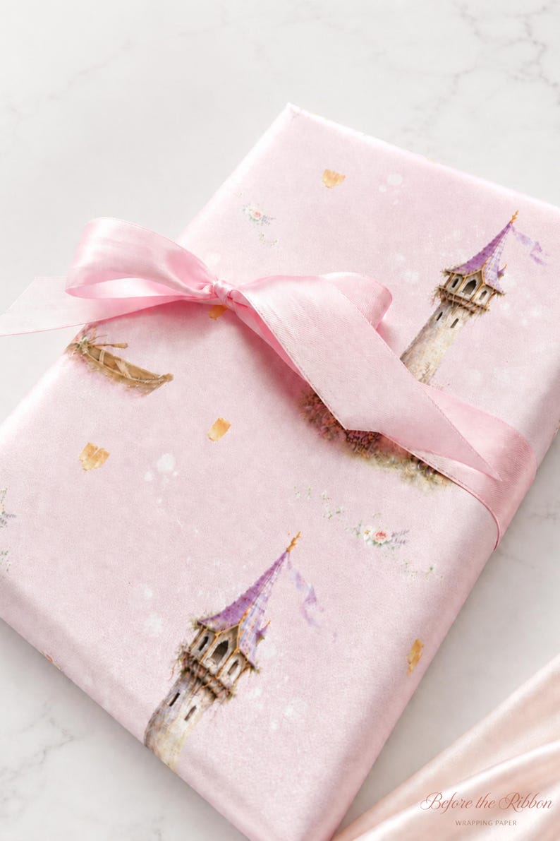 Puede incluir: Un regalo rectangular envuelto en papel rosa claro con un patr&oacute;n repetitivo de torres de piedra altas con techos c&oacute;nicos morados, botes peque&ntilde;os y detalles florales. Una cinta de sat&eacute;n rosa transl&uacute;cida est&aacute; atada en un lazo en la parte superior.