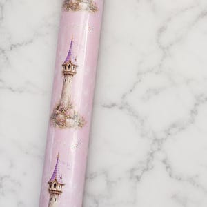 Puede incluir: Un rollo de papel de regalo rosa claro con un patr&oacute;n repetitivo de torres de piedra altas con techos c&oacute;nicos morados. Cada torre est&aacute; rodeada de peque&ntilde;as flores coloridas y vegetaci&oacute;n. El papel tiene un acabado liso y brillante.
