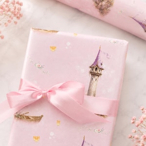 Puede incluir: Una caja de regalo rectangular envuelta en papel rosa claro con un patr&oacute;n repetitivo de torres de piedra altas con techos c&oacute;nicos morados, peque&ntilde;os botes de madera y detalles florales, atada con un lazo de cinta de sat&eacute;n rosa claro.