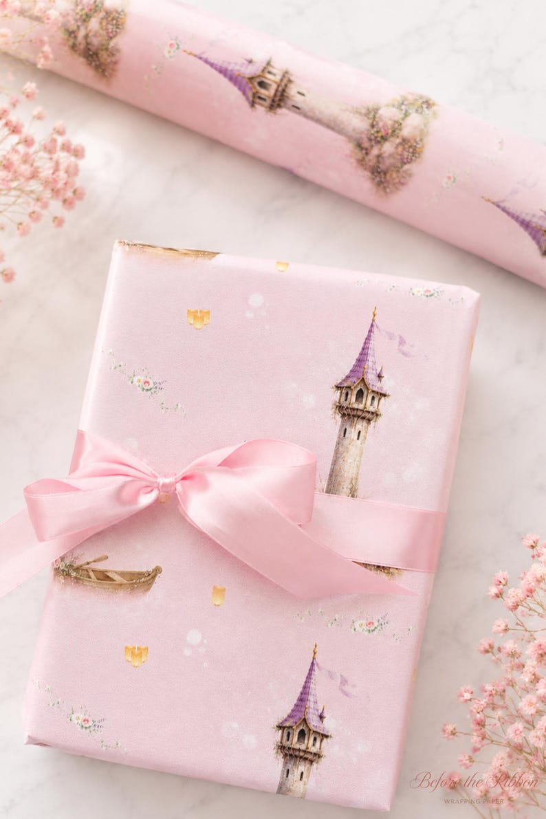 Puede incluir: Una caja de regalo rectangular envuelta en papel rosa claro con un patr&oacute;n repetitivo de torres de piedra altas con techos c&oacute;nicos morados, peque&ntilde;os botes de madera y detalles florales, atada con un lazo de cinta de sat&eacute;n rosa claro.