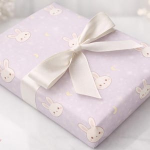 May include: A wrapped gift box featuring light purple wrapping paper with a repeating pattern of cartoon bunny faces and crescent moons. A cream-colored satin ribbon is tied in a bow on top. The gift is ready to be given.