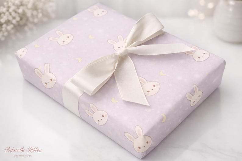 May include: A wrapped gift box featuring light purple wrapping paper with a repeating pattern of cartoon bunny faces and crescent moons. A cream-colored satin ribbon is tied in a bow on top. The gift is ready to be given.