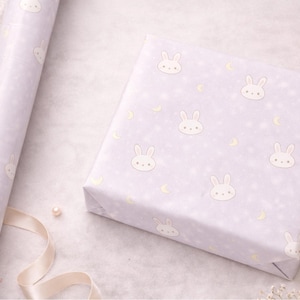 May include: A roll of wrapping paper and a wrapped gift box. Both are light purple with a repeating pattern of white bunny faces, stars, and crescent moons. The scene is decorated with pearls, flowers, and a ribbon.