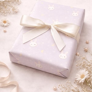 May include: A wrapped gift with light purple bunny-themed wrapping paper and a cream-colored satin ribbon. The paper has a repeating pattern of bunny faces and small yellow crescent moons. The bow is tied on top of the gift.