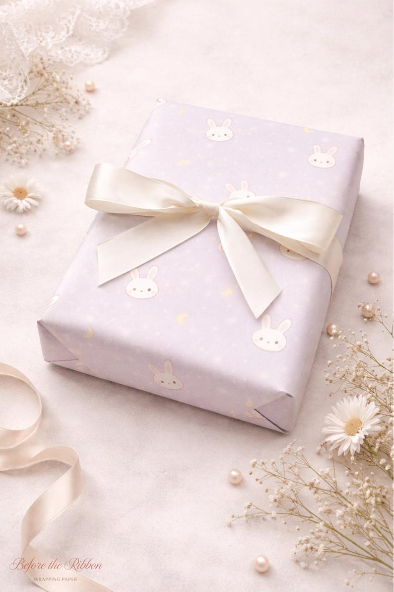 May include: A wrapped gift with light purple bunny-themed wrapping paper and a cream-colored satin ribbon. The paper has a repeating pattern of bunny faces and small yellow crescent moons. The bow is tied on top of the gift.