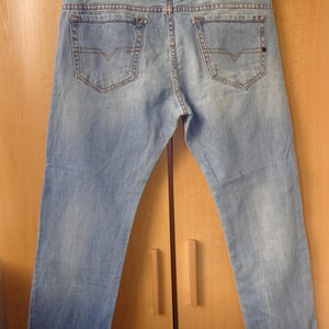 Könnte beinhalten: Hellblaue Jeans mit klassischem Fünf-Taschen-Design. Die Jeans haben eine ausgewaschene Optik und einen Lederaufnäher auf der Rückseite. Die Gesäßtaschen haben ein dekoratives Nahtdesign. Die Jeans hängen an einem schwarzen Kleiderbügel.