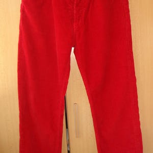 May include: Bright red corduroy pants featuring a button fly closure, five pockets, and silver-toned metal rivets. The trousers have a straight-leg cut and are displayed on a black hanger, showcasing the textured fabric and classic construction.