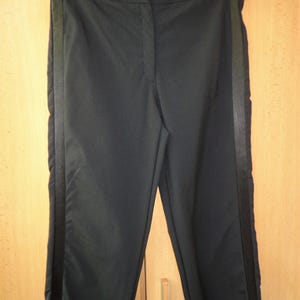 Puede incluir: Pantalones negros de corte recto con cierre de un solo botón. Los pantalones tienen una raya verde oscuro en el lateral de cada pierna. Colgados en una percha negra.