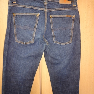 May include: Dark blue denim jeans featuring orange contrast stitching, two back pockets with decorative embroidery, a tan leather patch on the waistband with the text Nudie Jeans, and a black plastic hanger attached to the top.