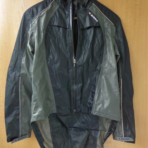May include: A black and grey athletic jacket with a full-length zip, hanging on a wooden hanger. The jacket has a high collar and features the text "X BIONIC" on the sleeve and shoulder. The jacket has a longer back hem.