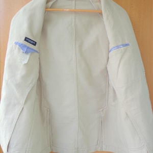 May include: A beige unlined blazer featuring a Gutteridge brand label on the interior chest pocket. The jacket includes two interior pockets with blue and white striped fabric trim, visible stitching along the seams, and a wooden hanger.