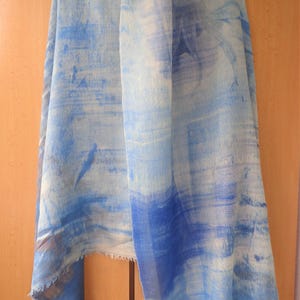 Peut inclure: Une écharpe rectangulaire présentant un motif abstrait bleu clair et blanc avec des textures rappelant des coups de pinceau horizontaux. Le tissu a un aspect doux et semi-transparent avec des bords effilochés le long de l'ourlet inférieur.