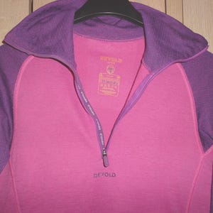 May include: A long-sleeve quarter-zip pullover featuring a bright pink torso and purple sleeves, collar, and trim. The garment includes a zip closure with the brand name Devold printed on the zip tape and the chest in a dark purple font.