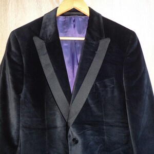 May include: A black velvet blazer with a satin lapel and a purple lining, hanging on a wooden hanger. The blazer has a single-button closure and a classic tailored design. The jacket is a formal wear item.