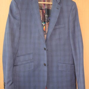 Op de afbeelding: Een blauwe blazer met ruitpatroon, voorzien van een reverskraag, twee knopen aan de voorzijde en twee klepzakken. De binnenvoering toont een gedetailleerde grafische print van een boekenplank vol boeken en een kleurrijke papegaai.