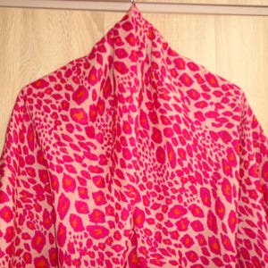 Peut inclure: Un vêtement rose et blanc à motifs, peut-être une chemise ou un chemisier, est suspendu à un cintre. Le tissu présente un motif léopard répétitif dans des tons de rose et d'orange. Le vêtement est fait d'une matière légère.