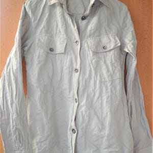 Pode incluir: Uma camisa de manga comprida cinza-claro com colarinho clássico, dois bolsos no peito com abas abotoadas e uma fileira central de botões. O tecido tem uma textura ligeiramente enrugada e mangas compridas com punhos abotoados.