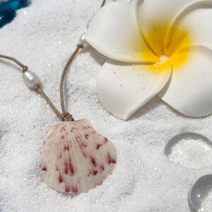 May include: A seashell pendant necklace with a pearl accent, displayed on white sand with a white and yellow flower. The shell is white with red markings. The necklace cord is brown.