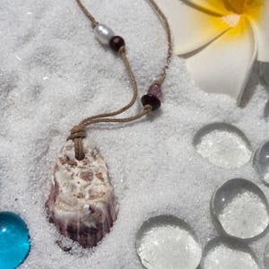 May include: A seashell pendant necklace with a natural cord. The pendant is a light purple and white seashell. The necklace has a pearl and dark red beads. The background is white sand with a white and yellow flower.