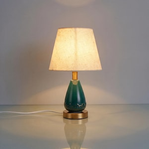 May include: A teal teardrop-shaped table lamp with a beige lampshade. The lamp has a gold-coloured base and accents. The lampshade is rectangular and the lamp is plugged into a white cord.