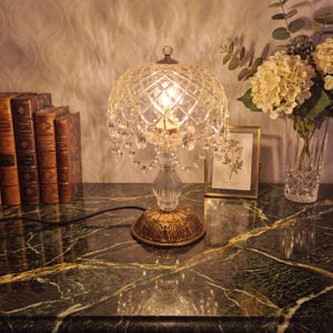 May include: A table lamp featuring a clear, faceted glass dome shade with hanging crystal droplets, a fluted glass stem, and a circular, ornate bronze-toned metal base with leaf-like engravings, emitting a warm glow from an internal light bulb.