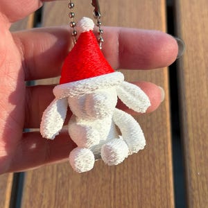 May include: A white 3D printed dog figurine keychain wearing a bright red Santa hat with a white pom-pom. The dog has floppy ears and a textured surface, attached to a silver-colored metal ball chain for hanging as a decorative ornament.