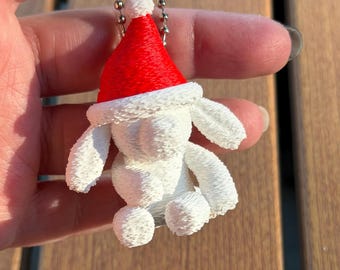May include: A white 3D printed dog figurine keychain wearing a bright red Santa hat with a white pom-pom. The dog has floppy ears and a textured surface, attached to a silver-colored metal ball chain for hanging as a decorative ornament.