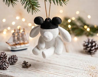 May include: A white plush animal ornament with long ears and a black cap featuring two round black ears on top. The ornament is suspended by a thin gold string from a green pine branch, showcasing a textured fabric surface and a seated posture.
