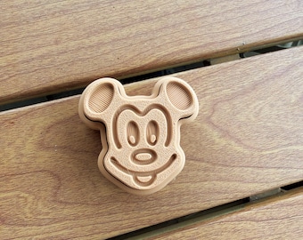 May include: A light tan, 3D-printed cookie cutter shaped like the head of a cartoon mouse with large rounded ears, a wide smiling mouth, and oval eyes. The surface of the plastic cutter shows fine horizontal layer lines from the printing process.