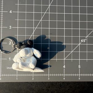 May include: A white and black dog-shaped keychain featuring a textured surface, attached to a silver metal split ring. The keychain is positioned on a gray cutting mat with white grid lines, showing a width of approximately 2.5 inches.