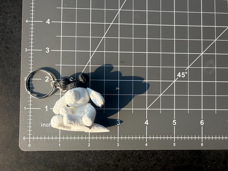 May include: A white and black dog-shaped keychain featuring a textured surface, attached to a silver metal split ring. The keychain is positioned on a gray cutting mat with white grid lines, showing a width of approximately 2.5 inches.