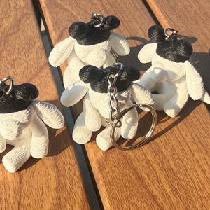 May include: Three small, textured sheep-shaped keychains featuring white bodies and black heads. Each figure has a metallic silver split ring attached to the top of the head, allowing for easy attachment to keys or bags. The surface has a distinct pattern.