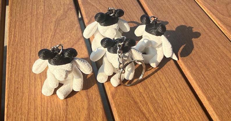 May include: Three small, textured sheep-shaped keychains featuring white bodies and black heads. Each figure has a metallic silver split ring attached to the top of the head, allowing for easy attachment to keys or bags. The surface has a distinct pattern.